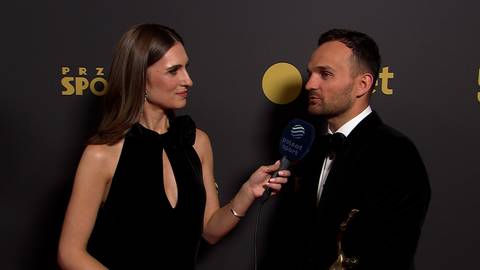 Bartosz Zmarzlik w czarnym smokingu udziela wywiadu kobiecie w czarnej sukni, trzymając w dłoni statuetkę.