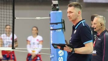 Trener siatk&oacute;wki z tabletem w ręku, patrzący w bok, z zawodniczkami w tle.