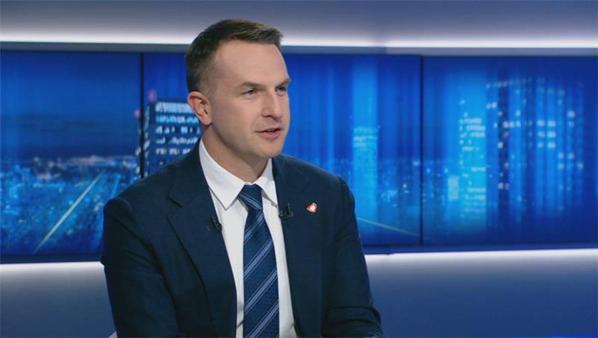 Mężczyzna w ciemnym garniturze i pasiastym krawacie, siedzący i mówiący, na tle ekranów z nocnym widokiem miasta.