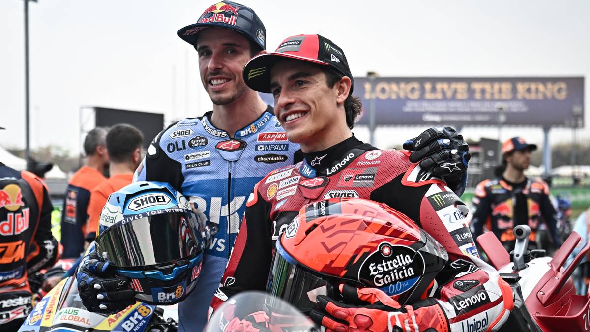 Dw&oacute;ch zawodnik&oacute;w MotoGP w pełnych strojach wyścigowych pozuje do zdjęcia, trzymając kaski motocyklowe. Jeden z nich to Marc Marquez w czerwono-białym stroju, drugi to Alex Marquez w niebiesko-białym stroju.