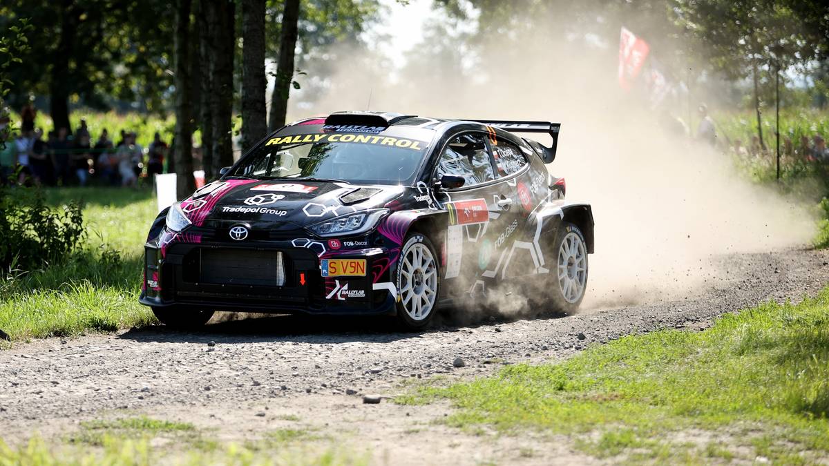 Samochód rajdowy Toyota Yaris GR Rally2 w locie, generujący tumany kurzu na szutrowej drodze, z widocznymi drzewami i widzami w tle.
