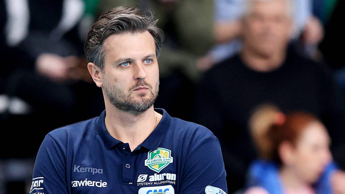 Trener siatk&oacute;wki w koszulce polo z herbem drużyny, z rękami skrzyżowanymi na piersi.