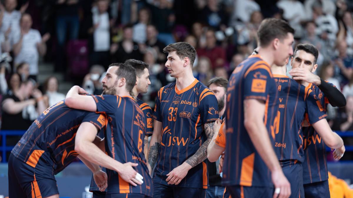 Siatkarze w strojach JSW Jastrzębski Węgiel świętują lub pocieszają się nawzajem na boisku, otoczeni przez kibic&oacute;w na trybunach.
