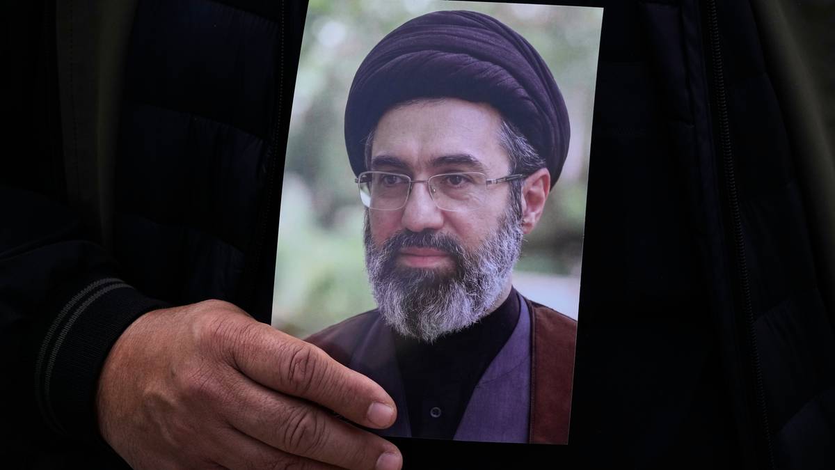 Zdjęcie przedstawia mężczyznę w czarnym nakryciu głowy i okularach, z brodą i wąsami. Trzyma je w dłoni osoba ubrana na czarno.