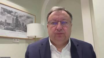 Mykoła Kniażycki, mężczyzna w średnim wieku w okularach i granatowej marynarce, siedzi przed ramą z obrazem przedstawiającym panoramę miasta.