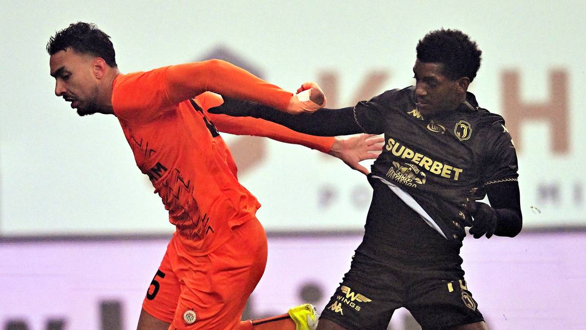 Dwóch piłkarzy na boisku piłkarskim, jeden w pomarańczowym stroju, drugi w czarnym, rywalizujący o piłkę.