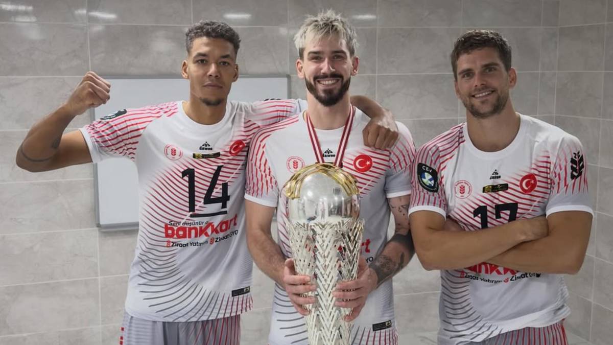 Trzech mężczyzn w białych koszulkach z czerwonymi elementami i numerami na plecach, z rękami na ramionach kolegi trzymającego wielkie, srebrne trofeum.