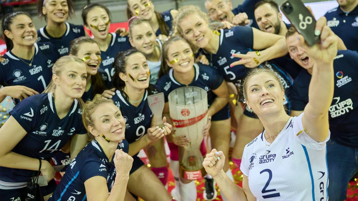 Grupa siatkarek świętuje zwycięstwo, jedna z nich robi selfie z pucharem.