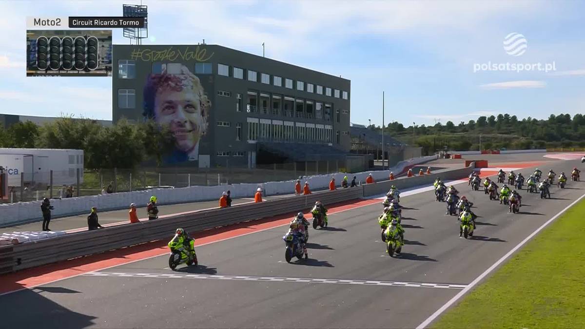 Wyścig motocyklowy Moto2 na torze, z zawodnikami na motocyklach i dużym portretem na budynku.