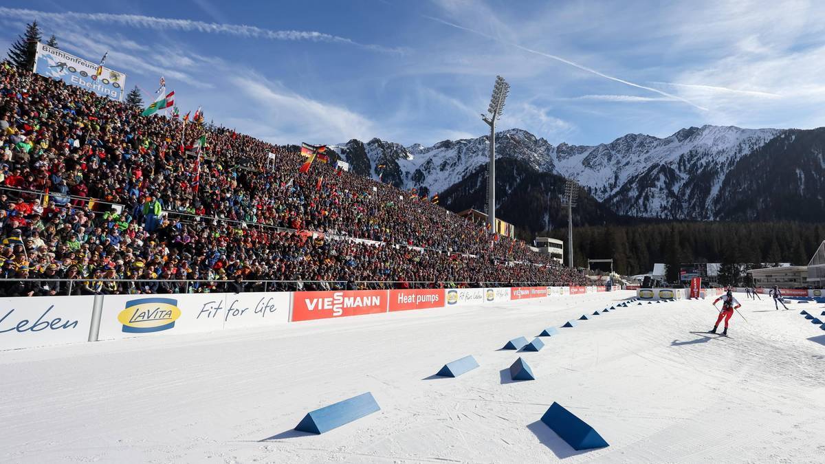 Widok na trasę biegu narciarskiego z trybunami wypełnionymi kibicami, w tle góry w śniegu. Jeden narciarz biegnie po trasie, a za nim ustawione są niebieskie stożki.
