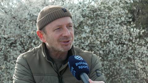 Mężczyzna w brązowej czapce i kurtce udziela wywiadu, trzymając mikrofon Polsat Sport na tle kwitnącego drzewa.