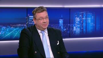 Mężczyzna w średnim wieku, ubrany w ciemny garnitur i jasny krawat, siedzi przed ekranami wyświetlającymi nocną panoramę miasta.