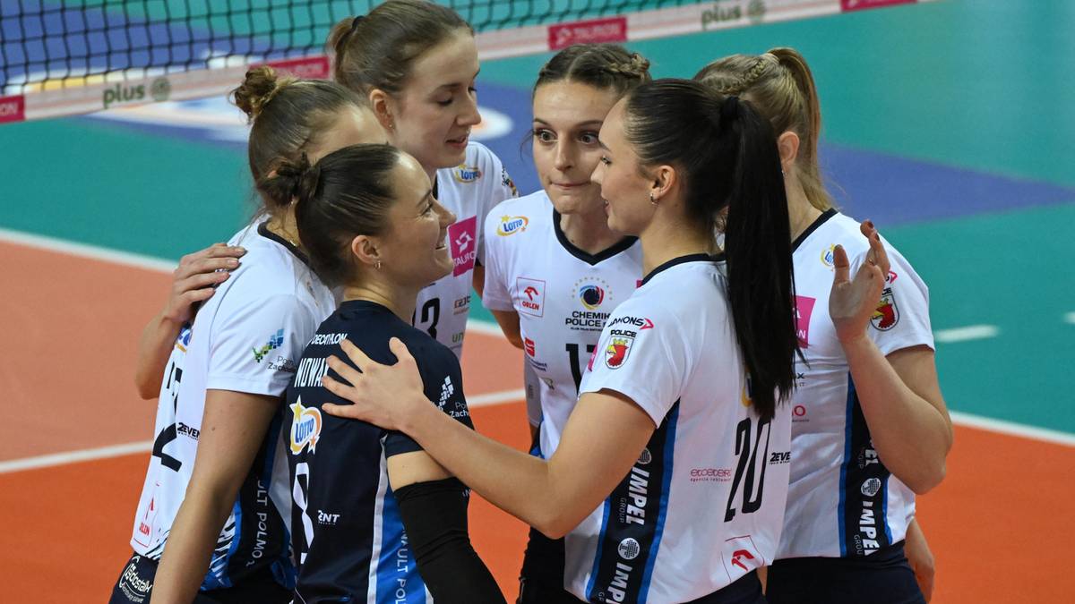 Grupa siatkarek w białych strojach sportowych otacza koleżankę w czarnym stroju, rozmawiając ze sobą na boisku do siatkówki.