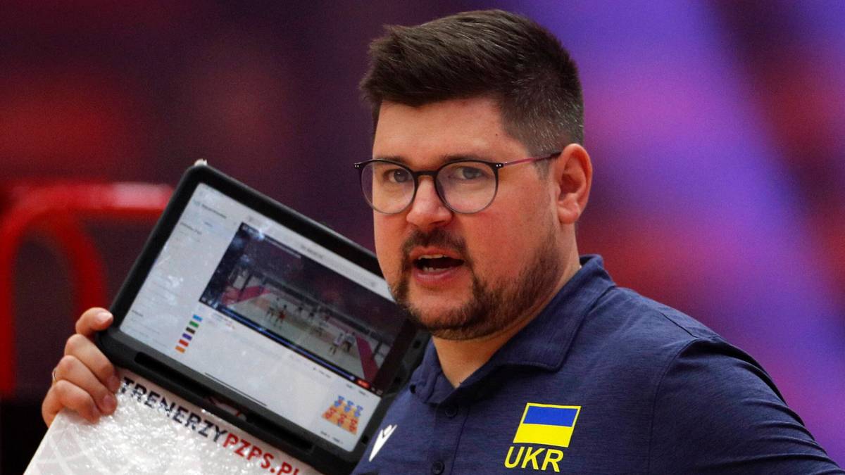 Trener z zarostem i okularami, ubrany w koszulkę z flagą Ukrainy, trzyma tablet z widokiem boiska do koszykówki.
