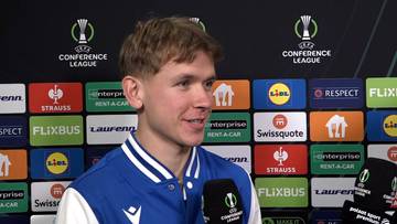 Młody mężczyzna udziela wywiadu na tle ścianki z logotypami Ligi Konferencji UEFA i sponsorów.