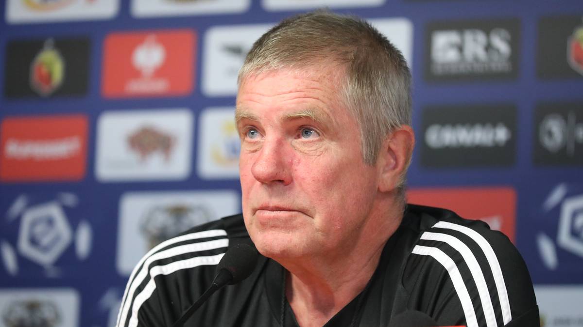 Portret starszego mężczyzny z krótkimi siwymi włosami, ubranego w czarną bluzę sportową z białymi pasami na ramionach, siedzącego przed ścianą z logotypami i panelami informacyjnymi.