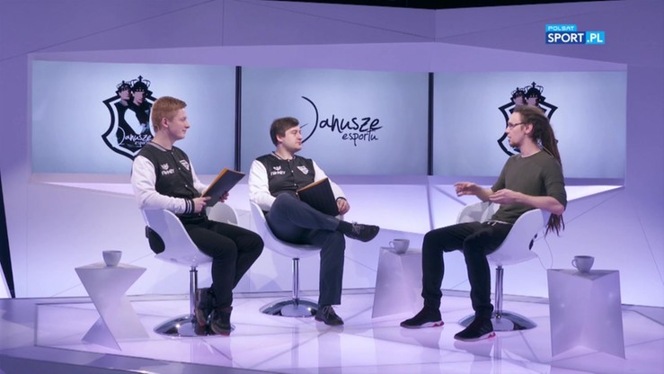 Trzech mężczyzn siedzących w studiu telewizyjnym, z logo "Janusze Esportu" na ekranie w tle.