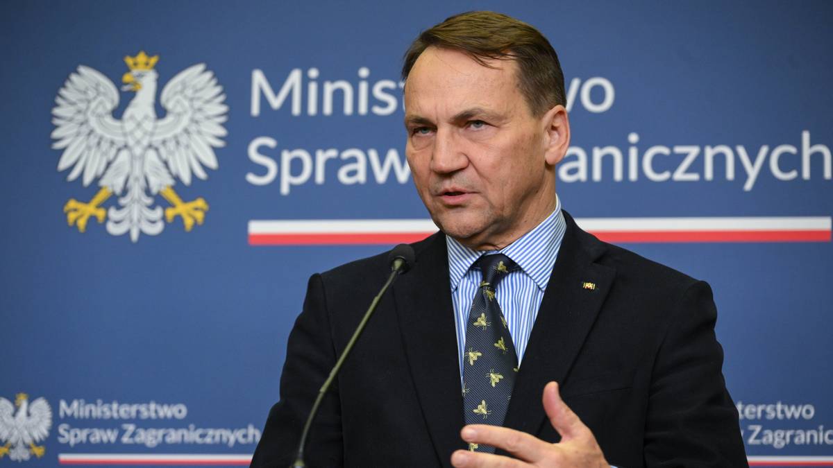 Radosław Sikorski przemawia przy mównicy na tle niebieskiej ściany z logo Ministerstwa Spraw Zagranicznych.
