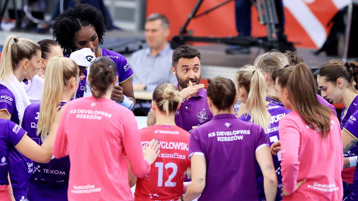 Drużyna siatkarska BKS Bostik ZGO Bielsko-Biała w różowych strojach zebrana wokół trenera, słuchająca jego wskazówek podczas meczu.