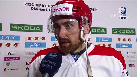 Hokeista w czerwonym kasku i białej koszulce z czerwonymi rękawami, z zarostem, patrzy w stronę kamery. Na jego piersi znajduje się mikrofon z logo "polsat sport". W tle widoczne są logotypy "SKODA" i "TAURON", a także napisy "2026 IIHF ICE HOCKEY WORLD CHAMPIONSHIP DIVISION I, GROUP B".