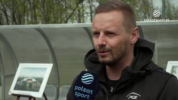 Mężczyzna w średnim wieku, z kr&oacute;tkimi włosami i kilkudniowym zarostem, ubrany w czarną bluzę z kapturem, udziela wywiadu. W tle widoczna jest ławka rezerwowych oraz dwa obrazy przedstawiające stadiony.