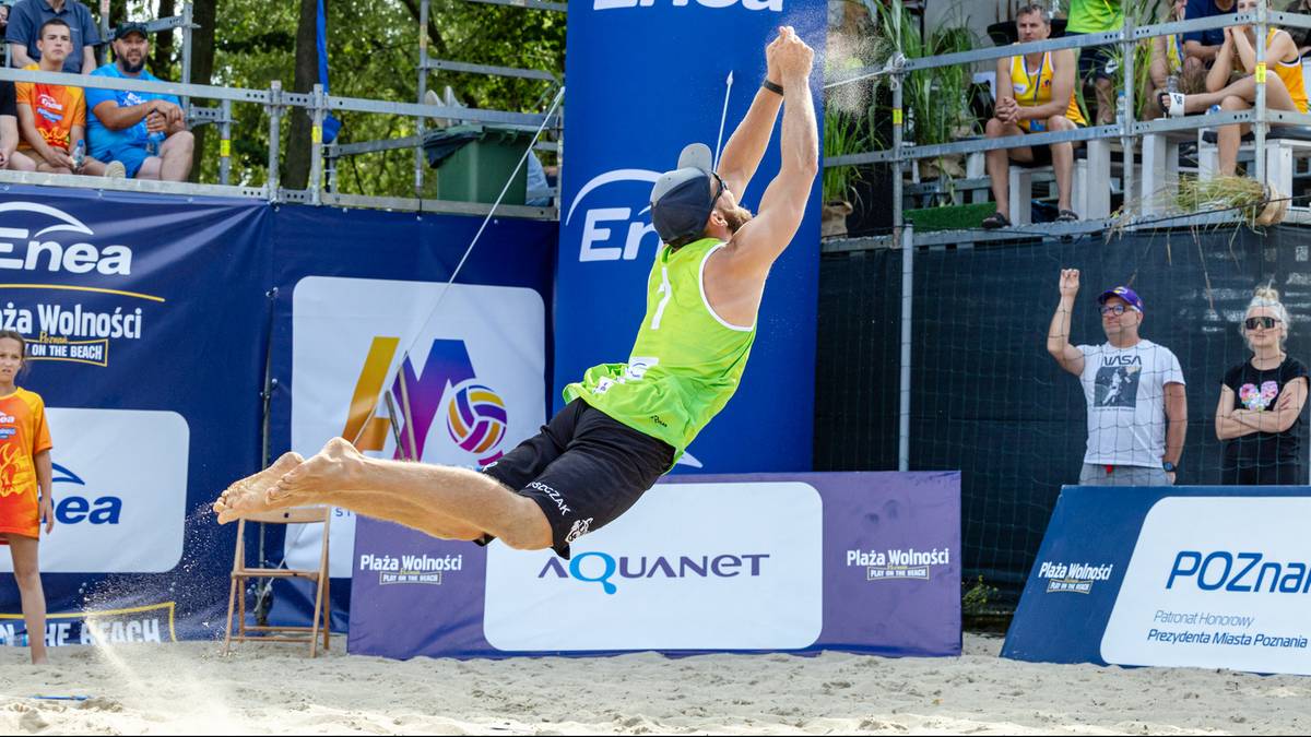 Siatkarz w trakcie akcji, wykonuje skok z wyciągniętymi rękami do góry. Ubrany jest w zieloną koszulkę i czarne spodenki.