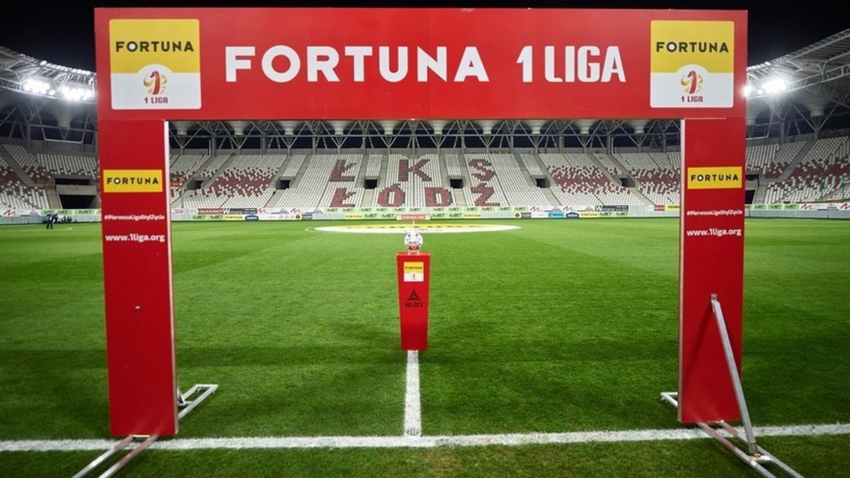 Czerwona brama z napisem Fortuna 1 Liga, ustawiona na murawie stadionu piłkarskiego. Za bramą widoczna jest trybuna z napisem "ŁKS Ł&Oacute;DŹ".