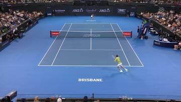 Gracz w ż&oacute;łtej koszulce odbija piłkę na niebieskim korcie tenisowym ATP Tour w Brisbane. W tle widoczni widzowie i inny gracz.