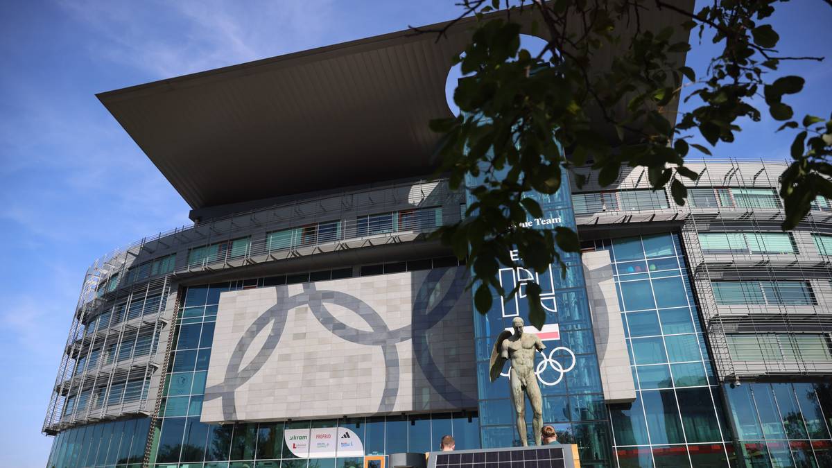 Nowoczesny stadion z przeszkloną fasadą i dachem, z symbolem k&oacute;ł olimpijskich na elewacji oraz statuą atlety.