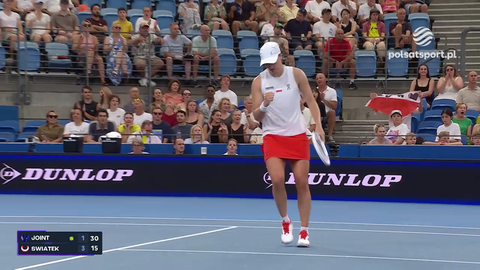 Iga Świątek, polska tenisistka, w białej koszulce i czerwonej sp&oacute;dniczce, z rakietą w dłoni, radośnie unosi pięść w geście zwycięstwa na tle trybun pełnych kibic&oacute;w.