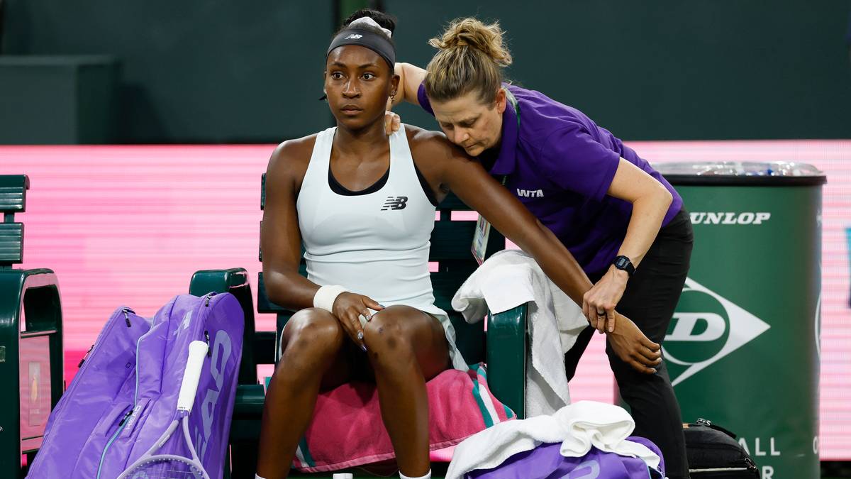 Coco Gauff siedzi na ławce tenisowej z bandażem na lewej ręce, podczas gdy fizjoterapeutka ją opatruje.