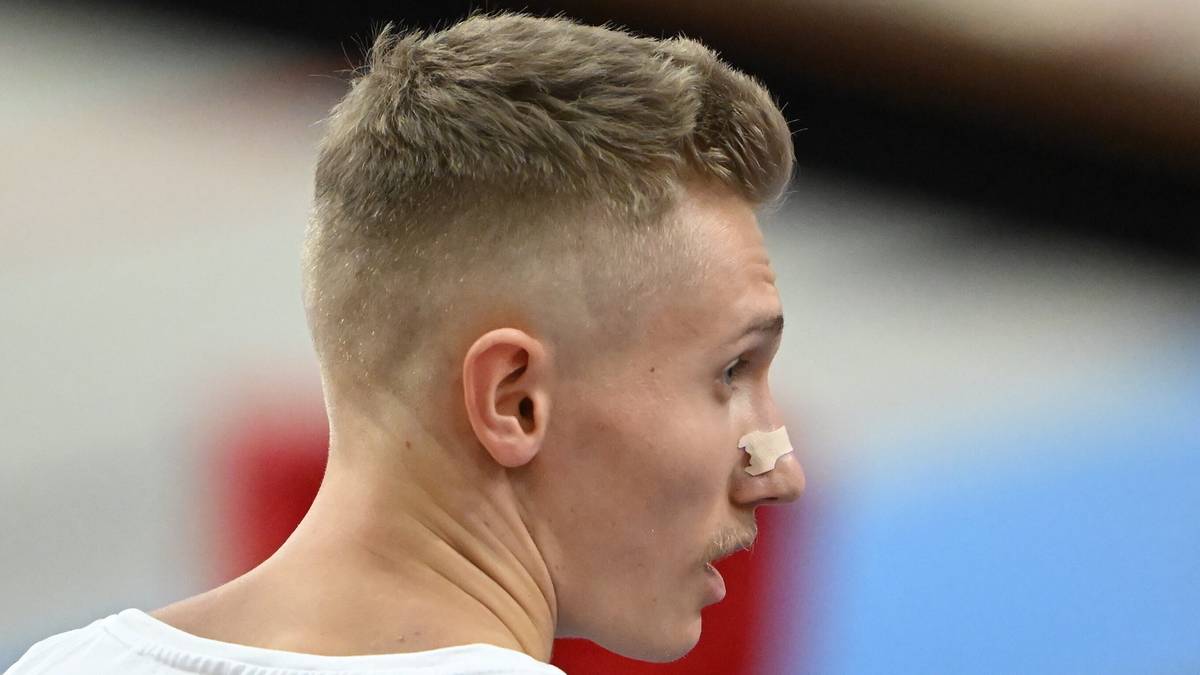 Mężczyzna z jasnymi, krótkimi włosami i plasterkiem na nosie, patrzący w bok z otwartymi ustami, w białej koszulce.