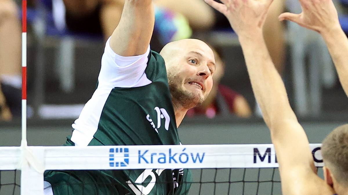 Siatkarz w białej koszulce z zielonymi akcentami wykonuje atak nad siatką podczas meczu siatkówki.