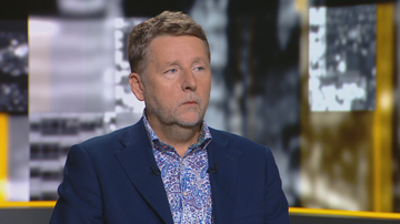 Mężczyzna w garniturze i wzorzystej koszuli siedzi przed kamerą z poważnym wyrazem twarzy.