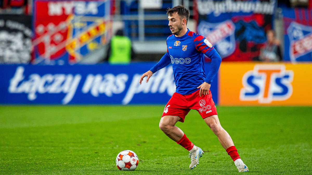 Piłkarz w niebiesko-czerwonym stroju prowadzi piłkę nożną na boisku.