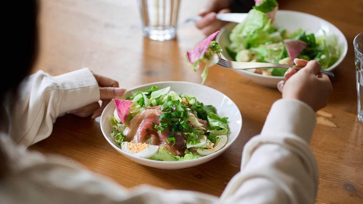 Dwie osoby jedzą sałatkę na drewnianym stole. Widok zza ramion os&oacute;b jedzących.