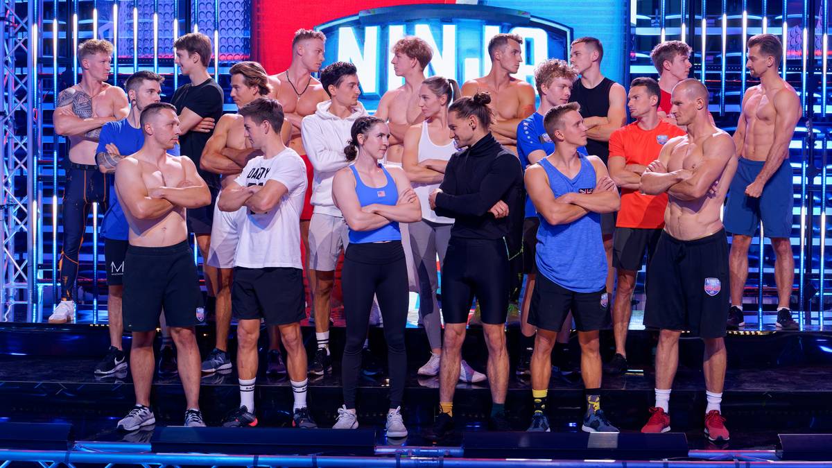 Grupa młodych, wysportowanych mężczyzn i dwie kobiety stoi na scenie, w tle widoczne są światła i logo "NINJA". Wszyscy ubrani są w sportowe stroje, niektórzy w koszulki z logo programu.