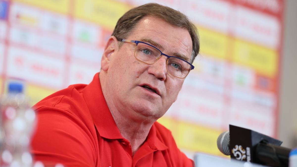 Mężczyzna w czerwonej koszulce z orłem na piersi, w okularach, mówi do mikrofonu podczas konferencji prasowej.