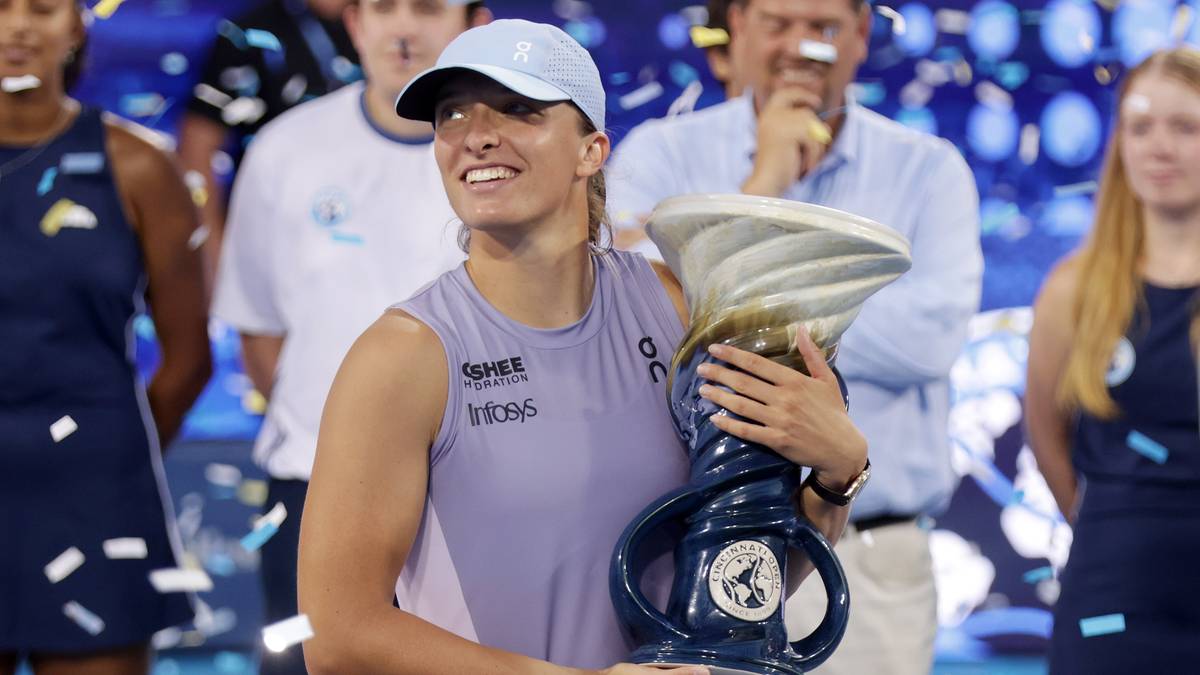Uśmiechnięta Iga Świątek trzyma w ramionach duże, faliste trofeum. W tle widać ludzi, a wokół spadają konfetti.