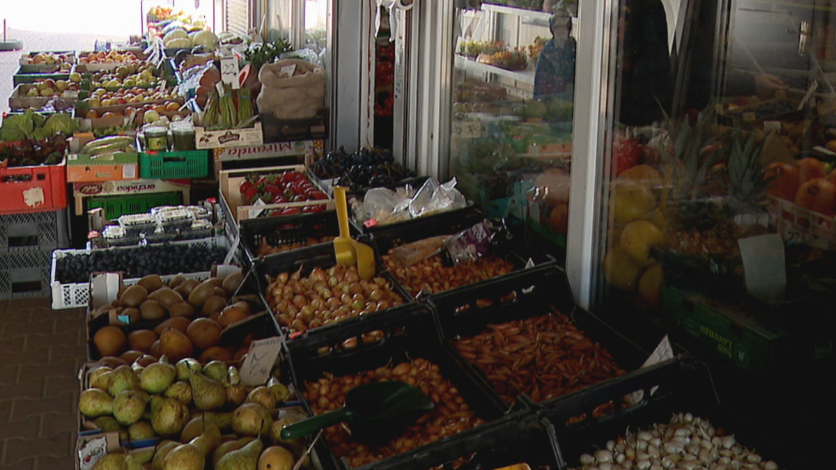 Zdjęcie przedstawia stoisko z r&oacute;żnorodnymi owocami i warzywami na targu, m.in. gruszki, cebule, winogrona, truskawki i inne produkty spożywcze.