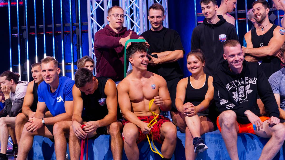 Grupa uczestnik&oacute;w programu sportowego pozuje do zdjęcia, niekt&oacute;rzy siedzą, inni stoją, uśmiechają się, w tle widać elementy scenografii.