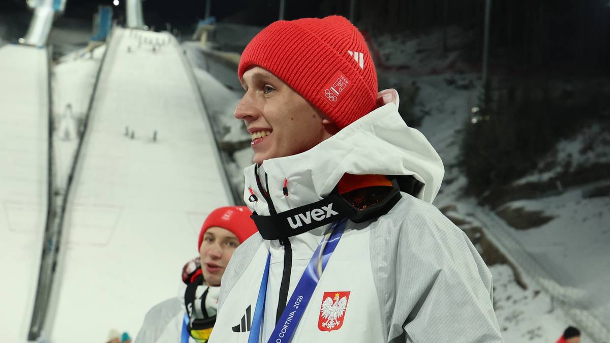 Skoczek narciarski w polskim stroju sportowym z medalem na szyi uśmiecha się, stojąc na tle skoczni narciarskiej nocą.