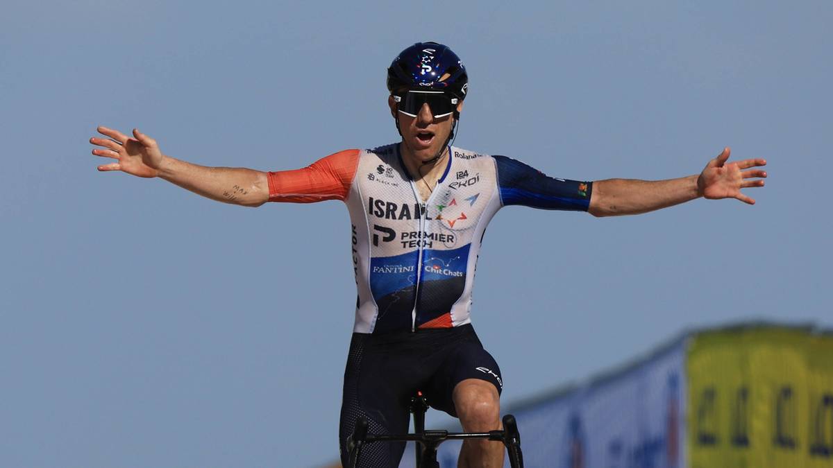 Kolarz w kombinezonie drużyny Israel Premier Tech z uniesionymi ramionami, na tle błękitnego nieba.