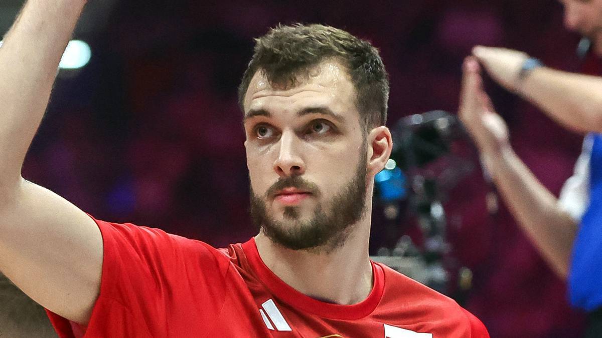 Mężczyzna w czerwonej koszulce z krótkim rękawem i krótkimi włosami, z brodą, patrzy w bok z lekko uniesioną ręką.