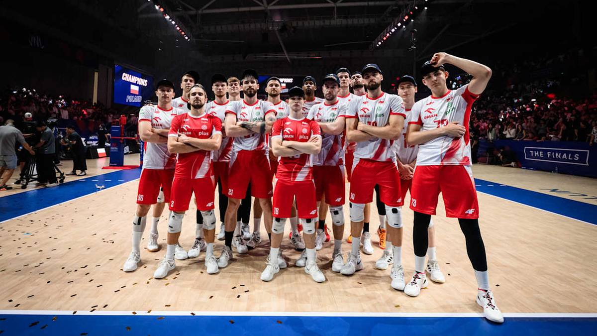 Drużyna polskich siatkarzy w strojach sportowych pozuje do zdjęcia na tle publiczności i trybun.