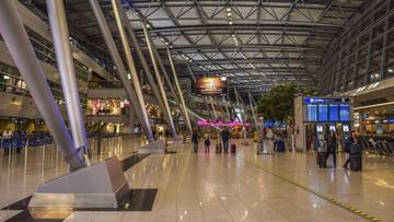 Wnętrze nowoczesnego lotniska z widocznymi stalowymi konstrukcjami i tablicami informacyjnymi.