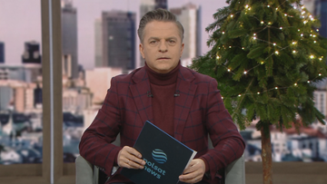 Mężczyzna w garniturze, trzymający w rękach dokument z logo Polsat News, siedzi na kanapie w studiu telewizyjnym z choinką w tle.