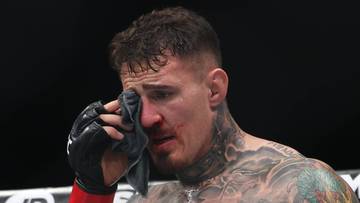 Tom Aspinall, zawodnik MMA, ociera twarz ręcznikiem, ubrany w spodenki UFC i rękawice bokserskie.