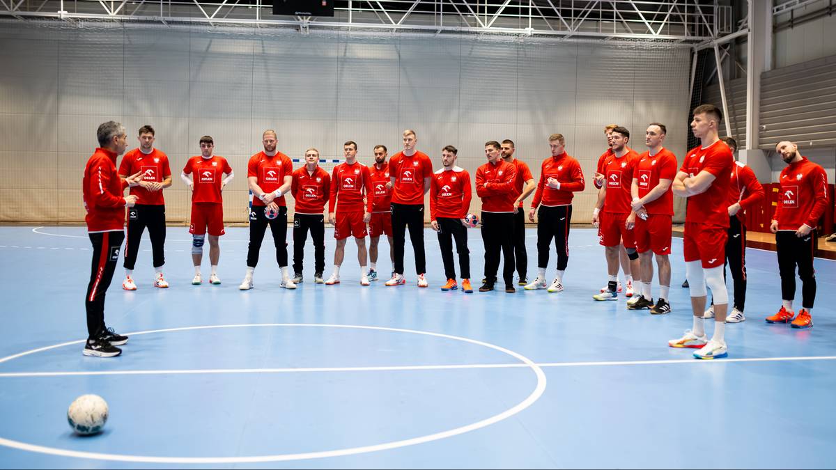 Drużyna piłkarzy ręcznych w czerwonych strojach treningowych stoi na hali sportowej, zebrana wok&oacute;ł trenera.
