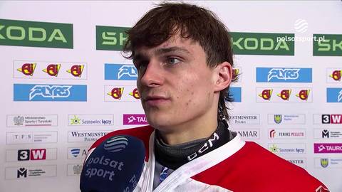Portret młodego mężczyzny w czerwonej bluzie sportowej, trzymającego mikrofon. Tło stanowi ściana z logotypami sponsor&oacute;w.